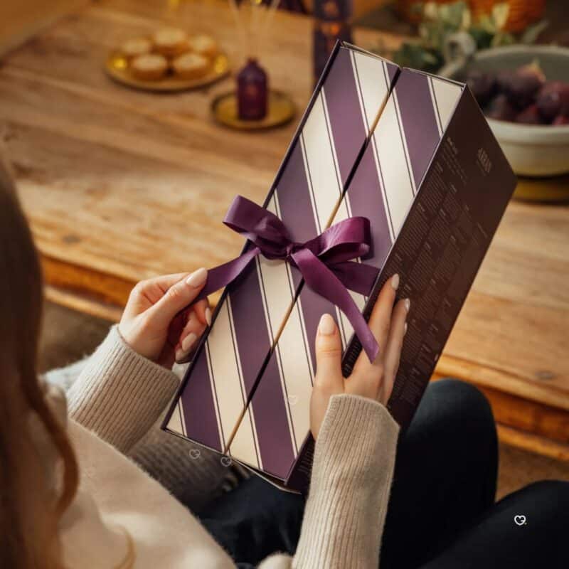Hands holding the ARRAN Advent Calendar 2026 tied with a purple ribbon on a festive table